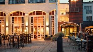 An outdoor dining area with tables and chairs, a building with arched windows, and a flag on a pole. - Bilderberg Résidence Groot Heideborgh (Garderen)