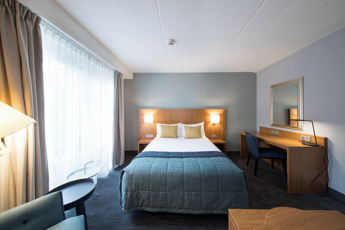 Habitación de hotel con cama, escritorio, silla y un gran ventanal con cortinas.