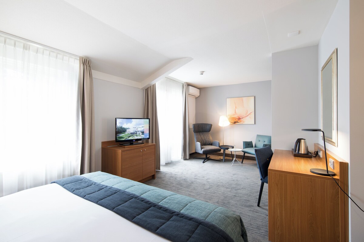 A hotel room with a bed, a television, a desk with a lamp, a chair, and a desk lamp.