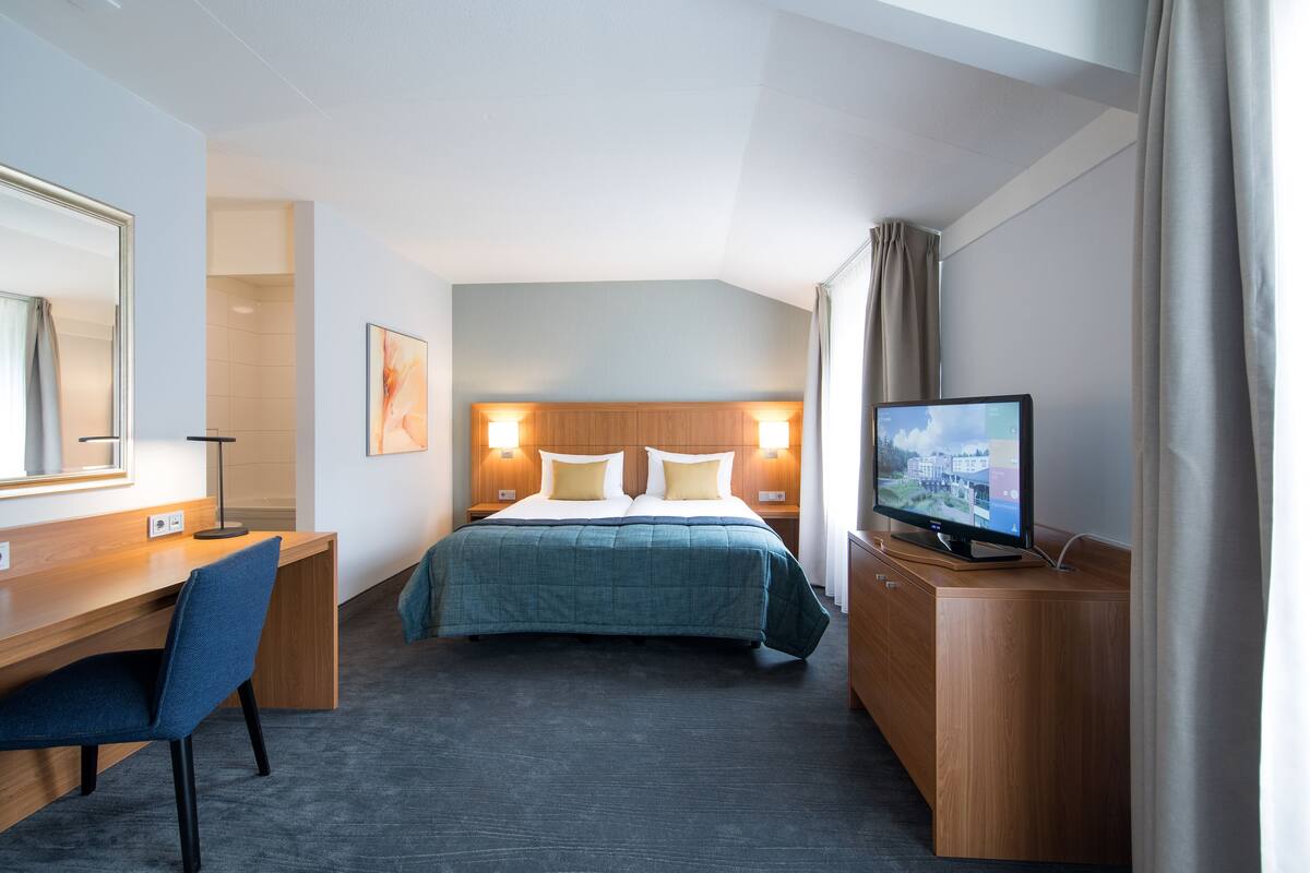 Habitación de hotel con cama, escritorio, silla, televisión y un amplio ventanal con cortinas.