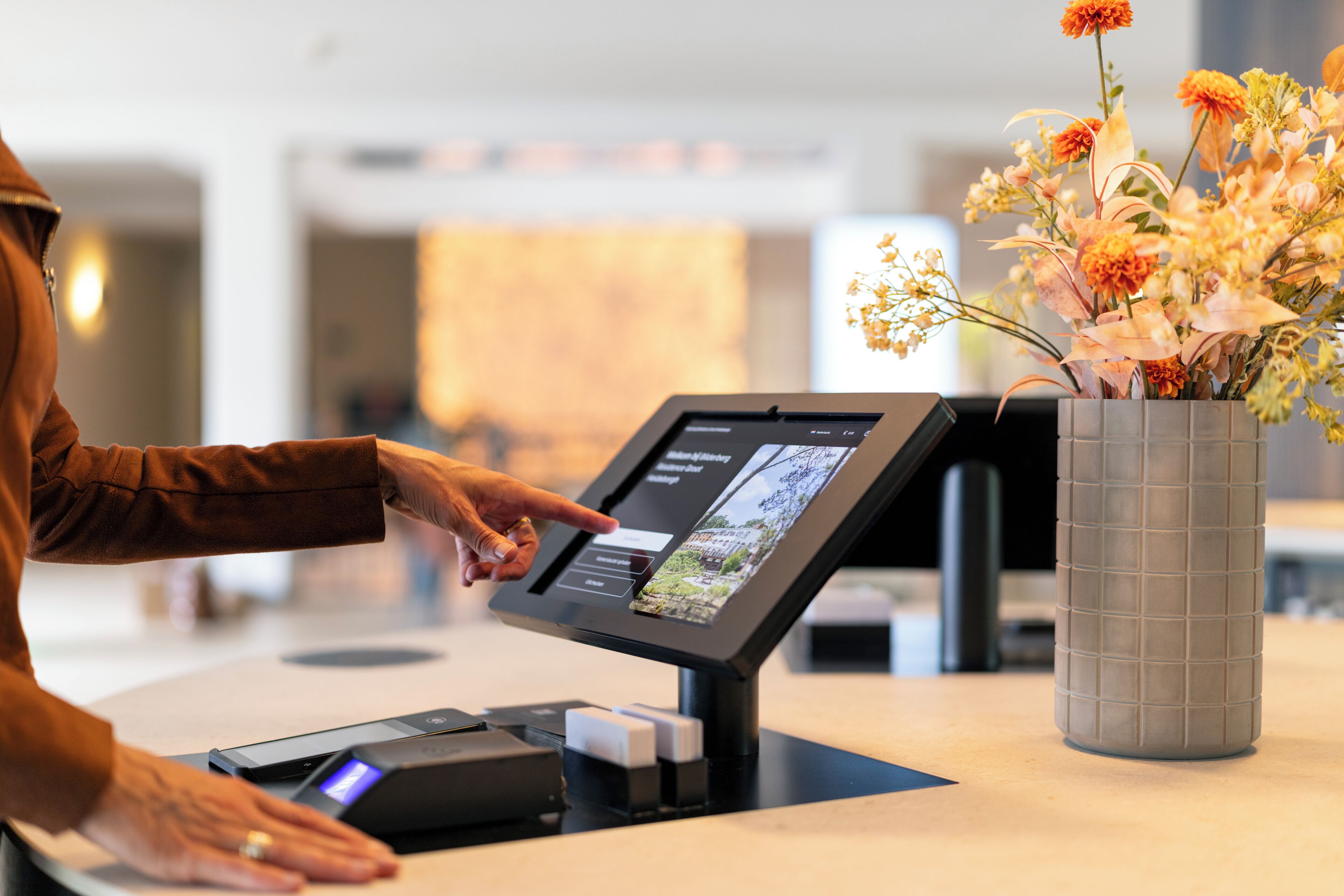 Eine Person bedient einen digitalen Kiosk in einem Hotel, an der Rezeption steht ein Blumenstrauß.