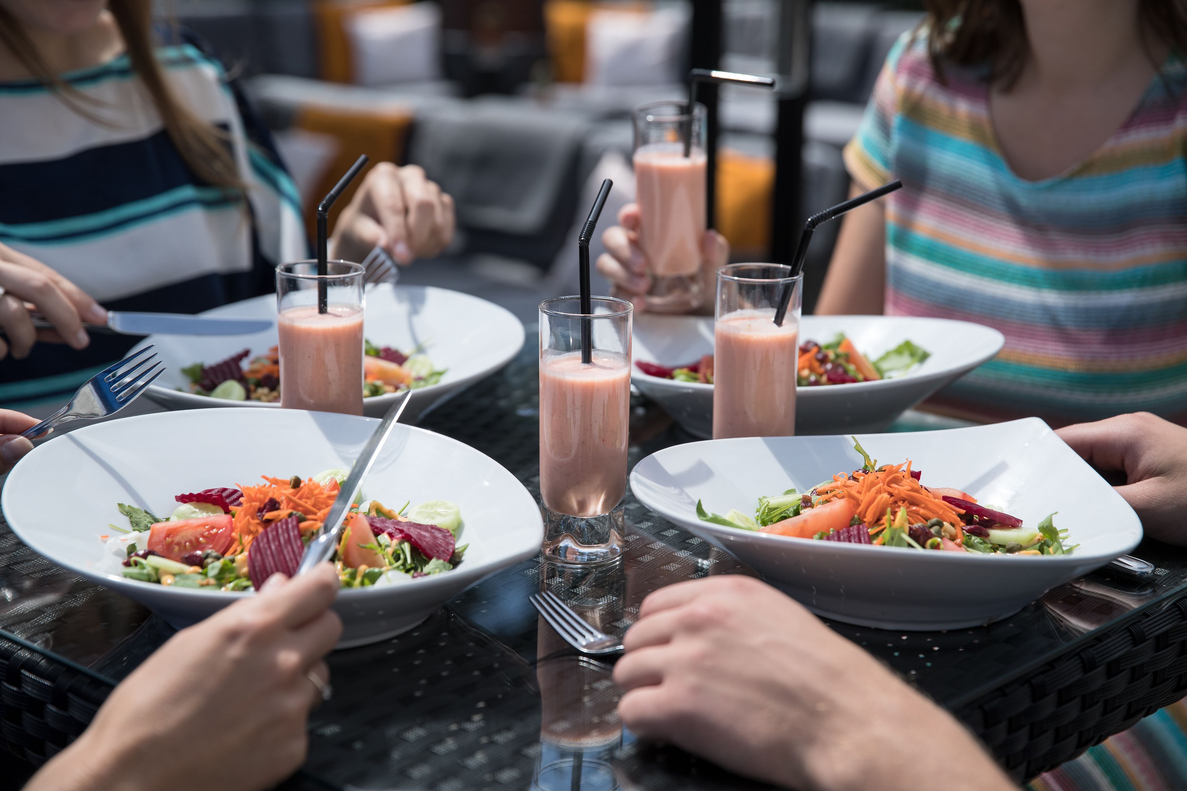 Een groep mensen dineert in een openluchtrestaurant, met witte borden vol salade en roze smoothies.