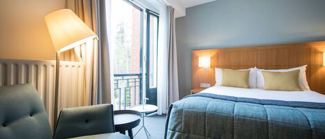 A hotel room with a bed, a chair, a small table, and a view of the outdoors.