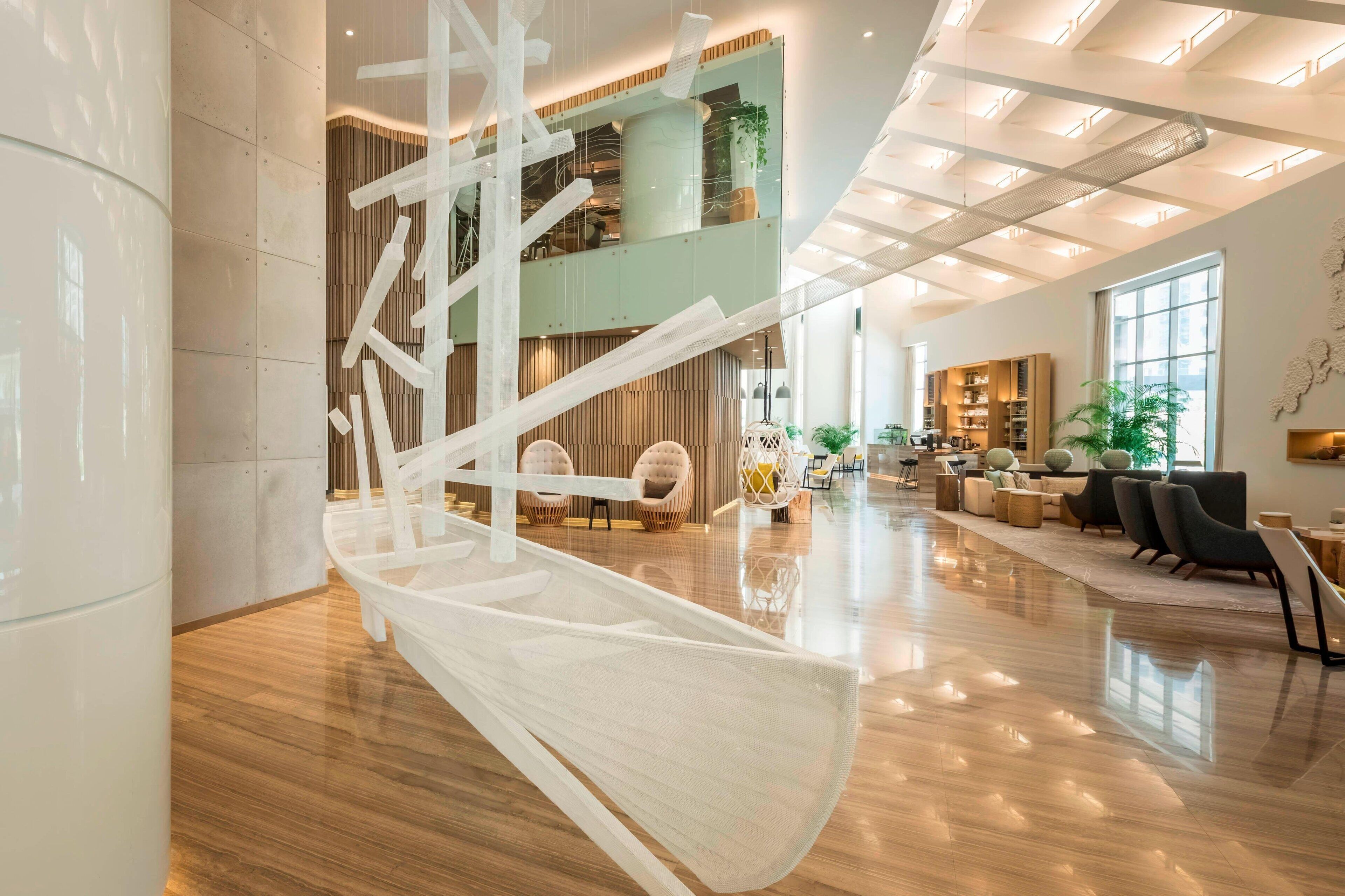 A spacious lobby with a modern staircase, seating areas, and a second-floor balcony with plants.