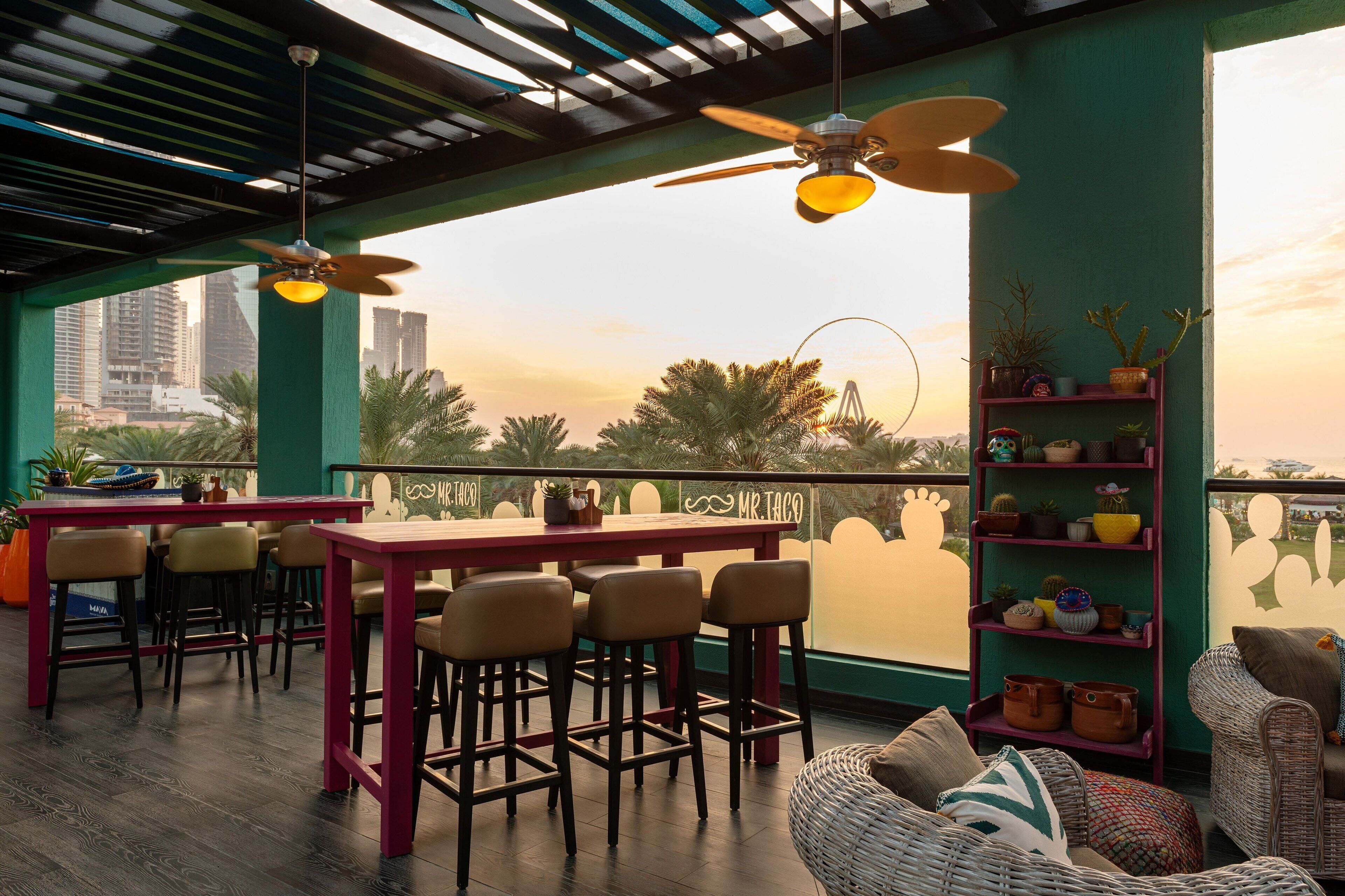 Un bar situé sur le toit, offrant une vue sur les gratte-ciel de la ville, avec une table et des chaises, un ventilateur de plafond et une étagère ornée de plantes.