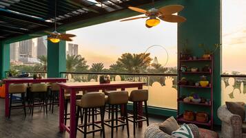 Un bar situé sur le toit, offrant une vue sur les gratte-ciel de la ville, avec une table et des chaises, un ventilateur de plafond et une étagère ornée de plantes.