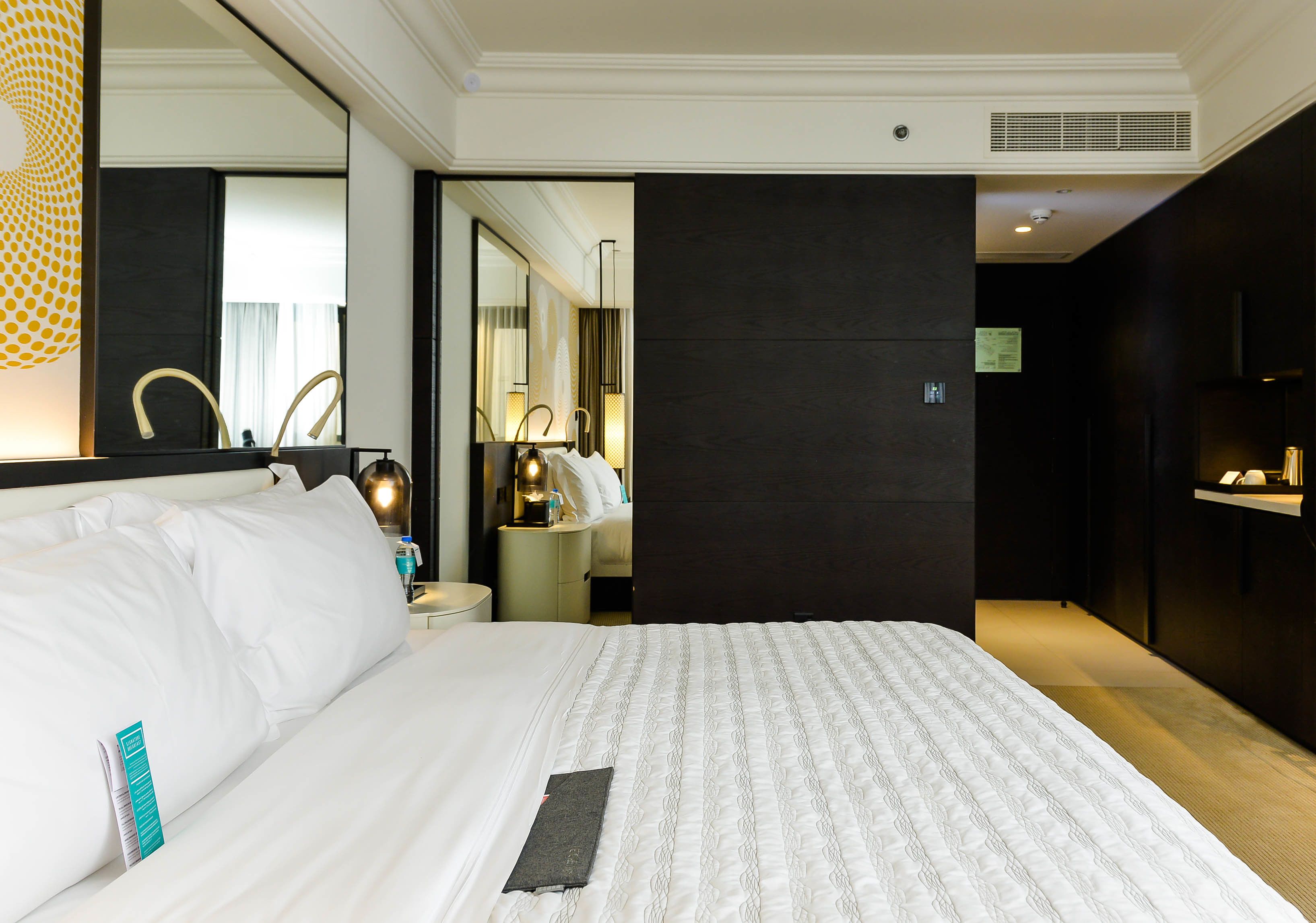 Une chambre d’hôtel avec un grand lit, une table de chevet avec une lampe, un miroir et un mur de couleur sombre.
