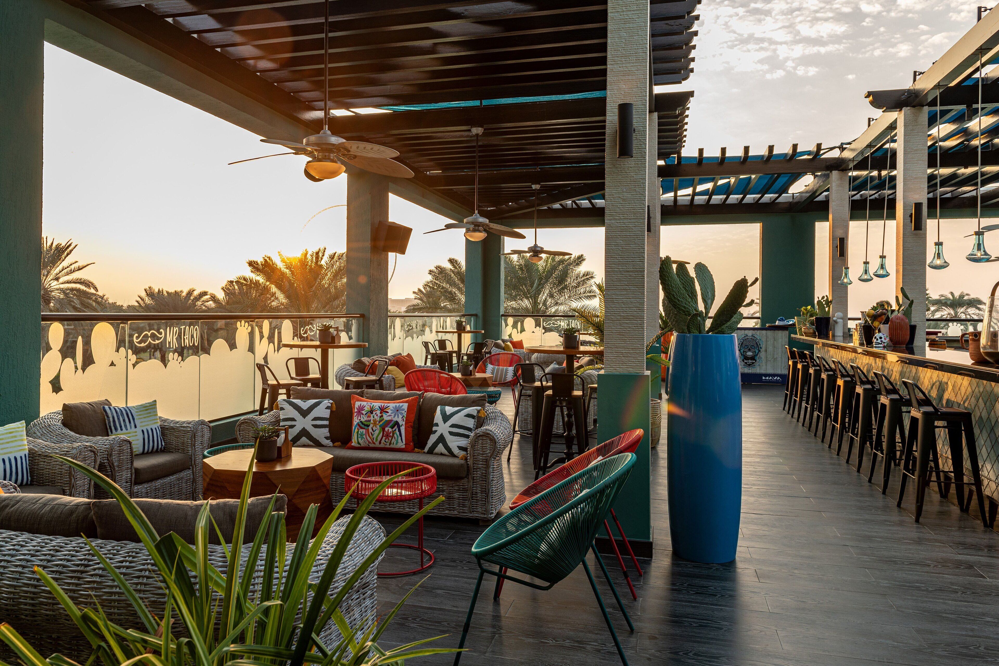 A rooftop bar with wicker furniture, ceiling fans, and a view of palm trees.