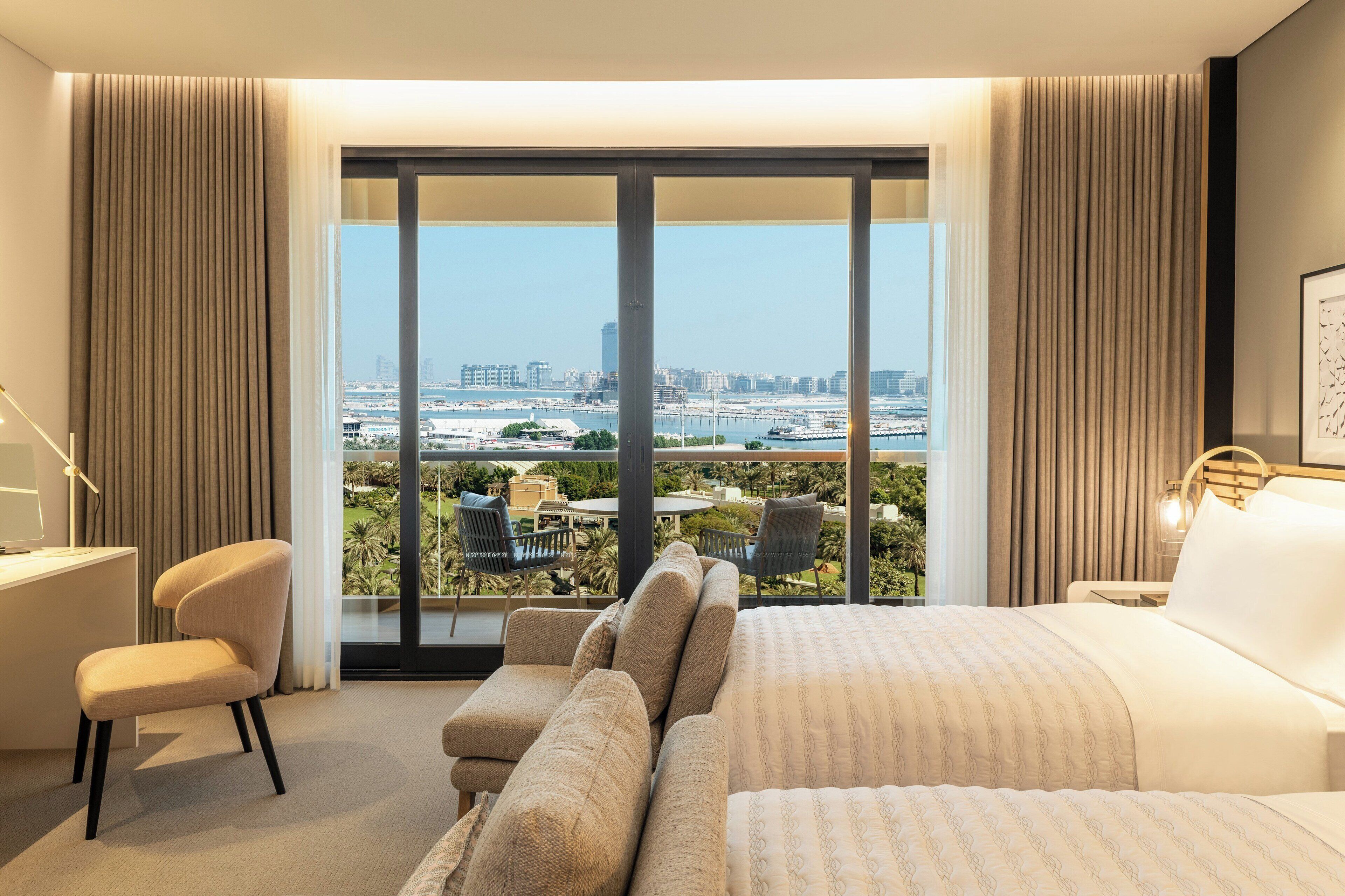 A hotel room with a large bed, a desk, and a view of a cityscape through floor-to-ceiling windows.