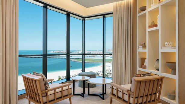 Ein Zimmer mit großen Fenstern, die auf Strand und Stadtlandschaft blicken, ausgestattet mit zwei Stühlen und einem kleinen Tisch.