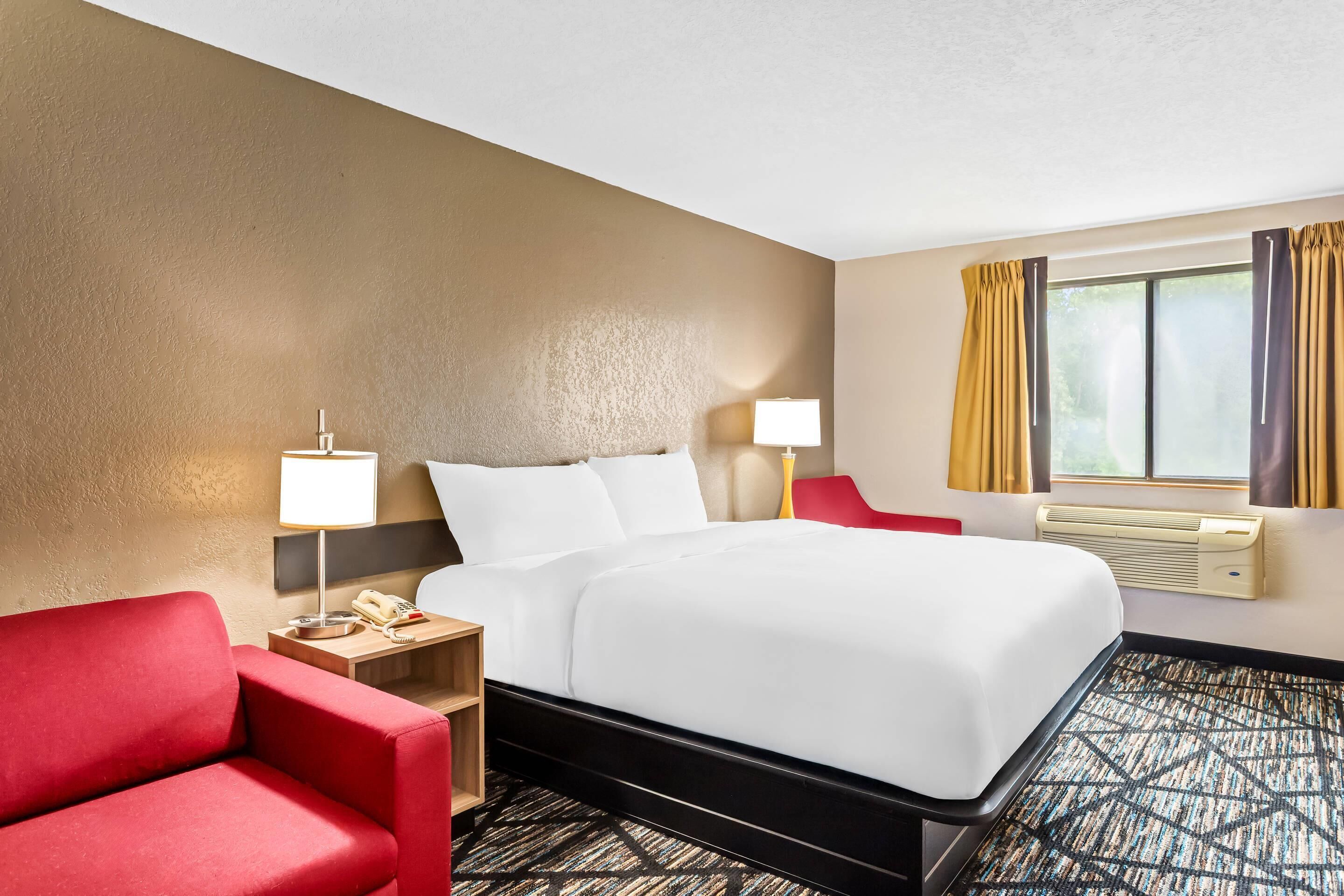A hotel room with a bed, a red armchair, a desk with a lamp, and a window with curtains.