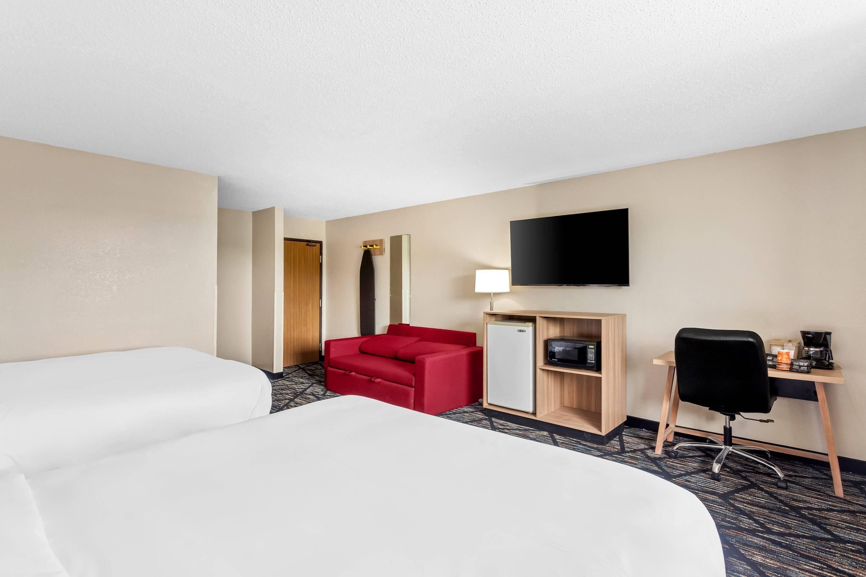 A hotel room with two beds, a red sofa, a desk with a computer, a TV, and a refrigerator.