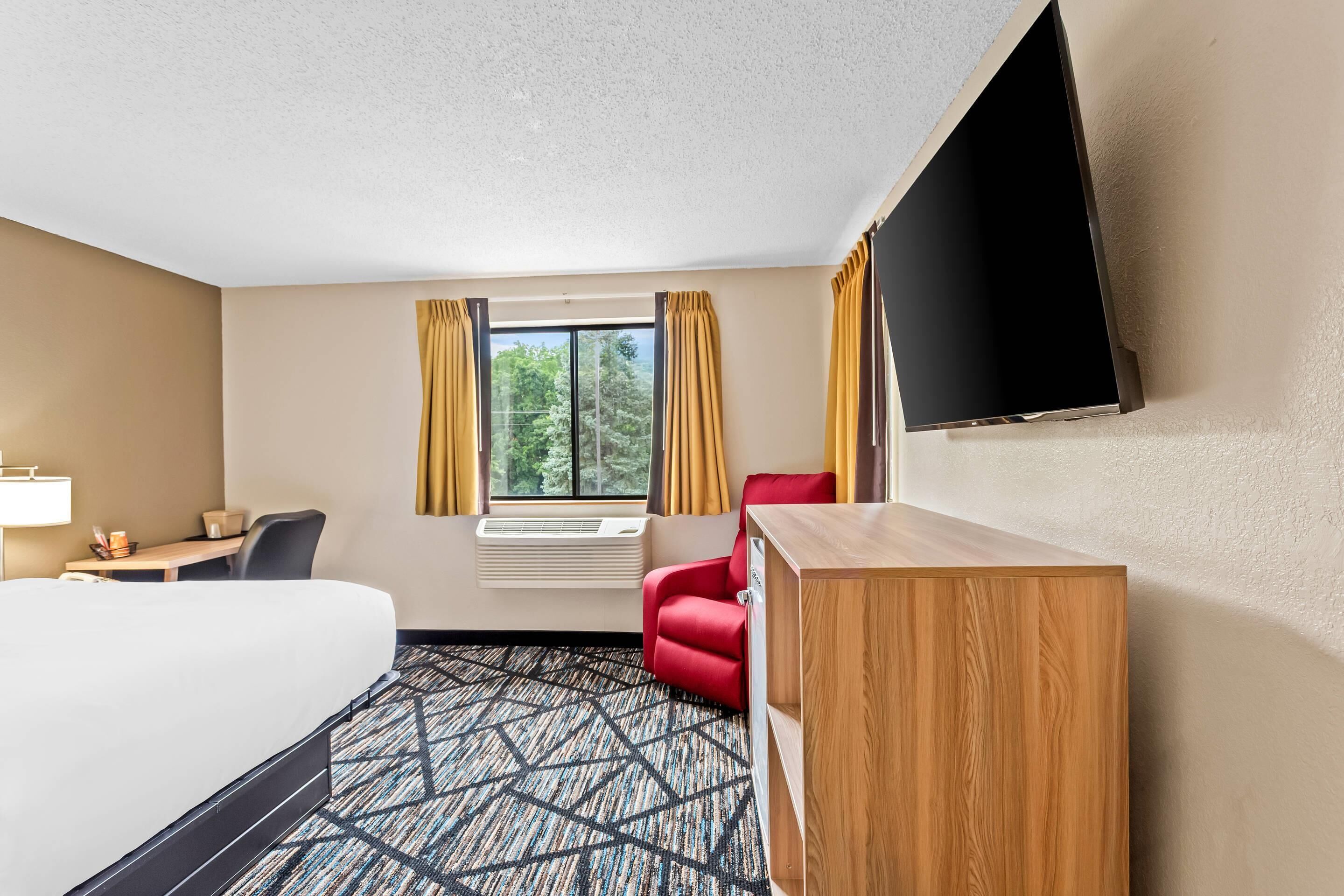 Quarto de hotel com uma cama grande, uma escrivaninha com cadeira, uma poltrona vermelha, um armário de madeira e uma televisão de tela plana na parede.