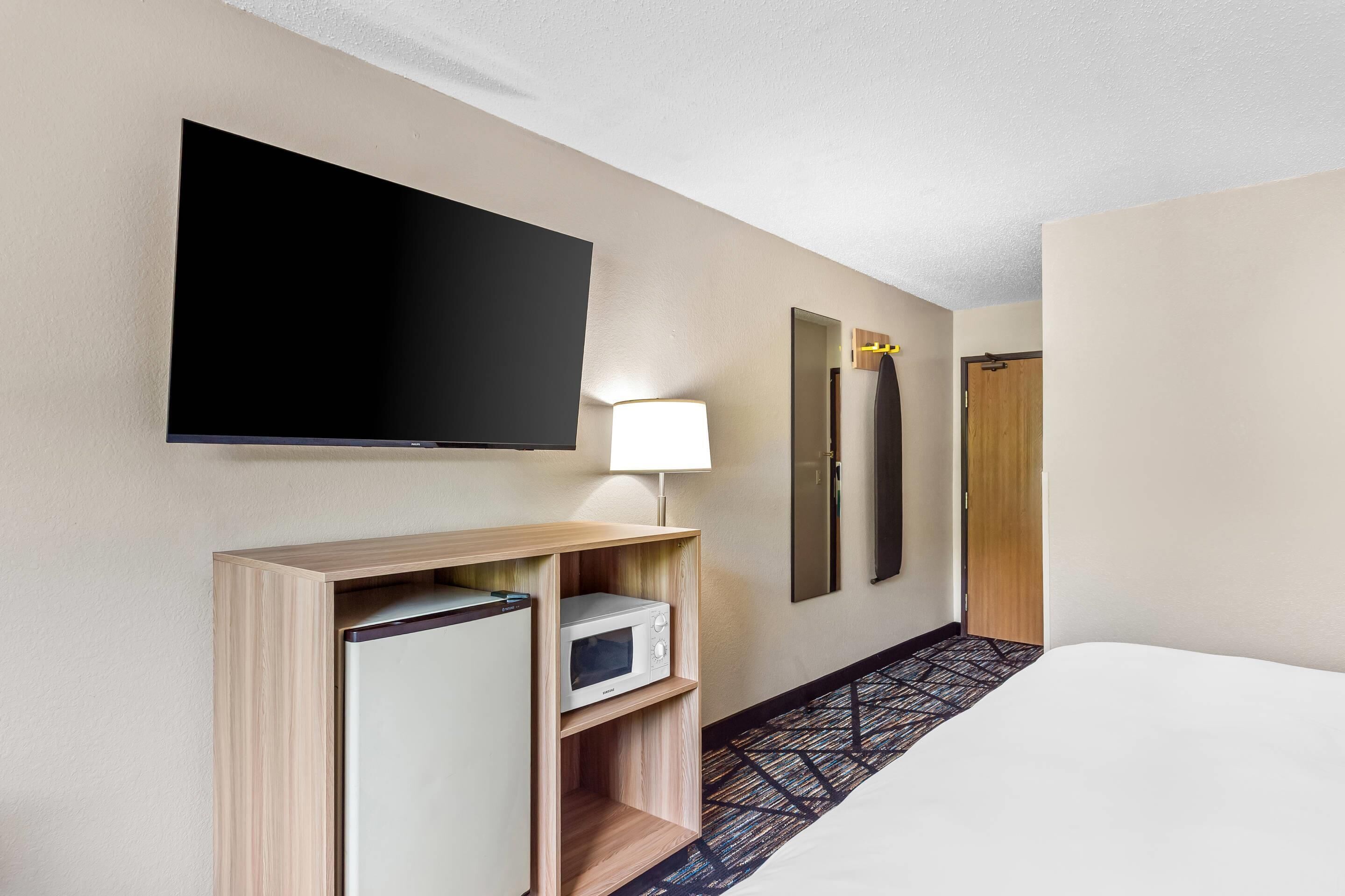 A hotel room with a bed, a flat-screen TV mounted on the wall, a microwave, a lamp, and a door.