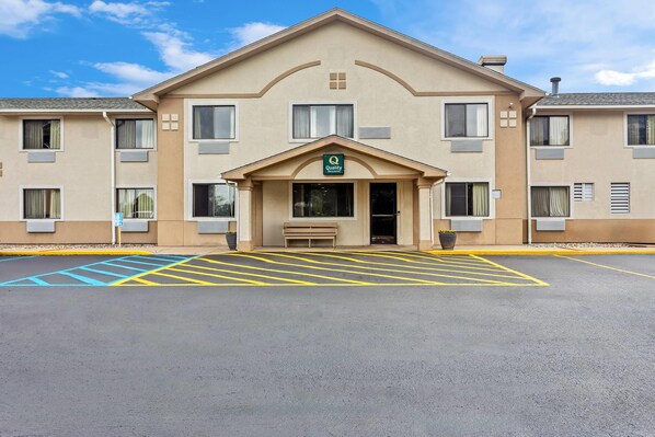Hotel com fachada bege, entrada central com uma placa 'Quality' e estacionamento com marcações amarelas e azuis.