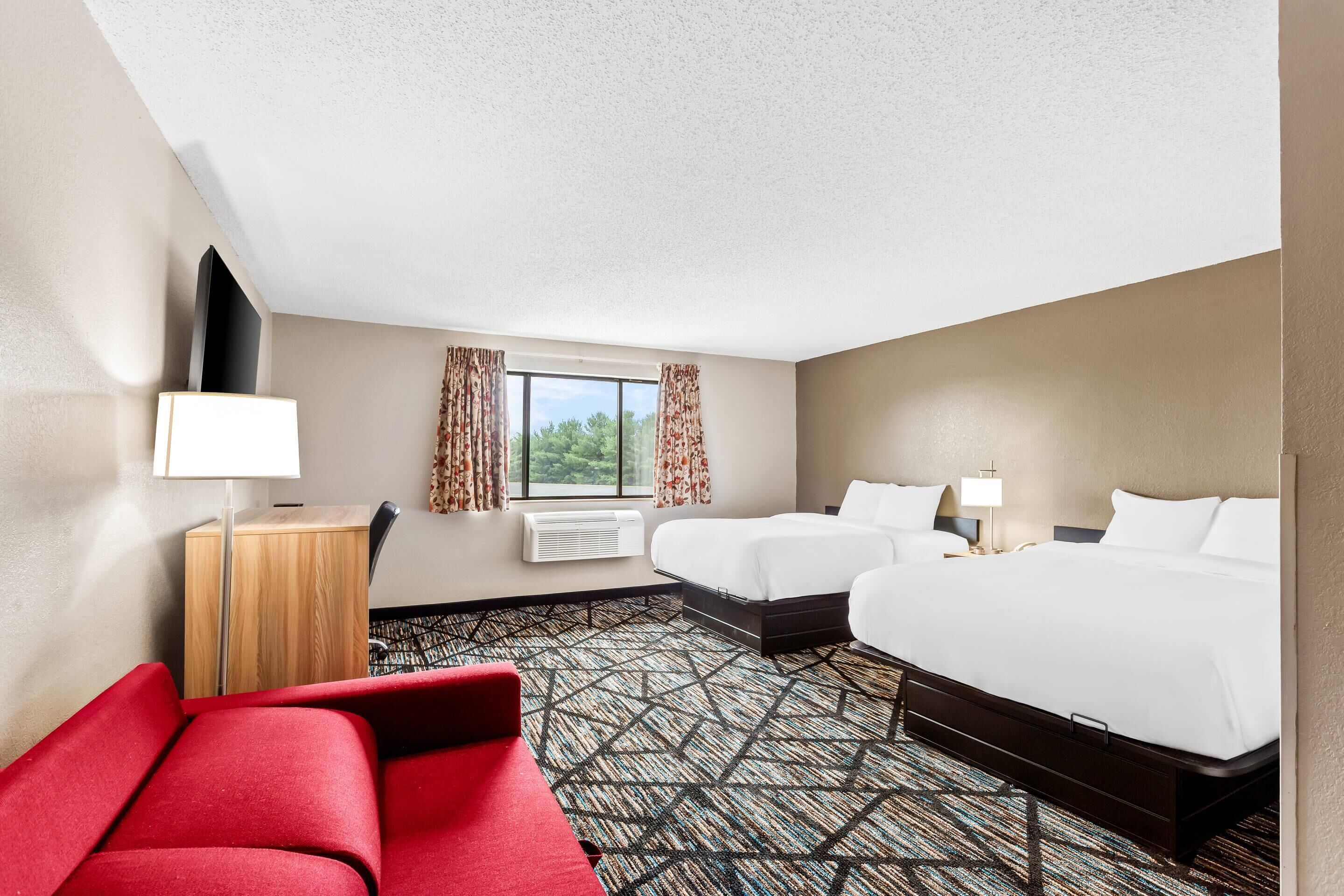 Habitación de hotel con dos camas, un sofá rojo, un televisor de pantalla plana, un escritorio y una ventana con cortinas.