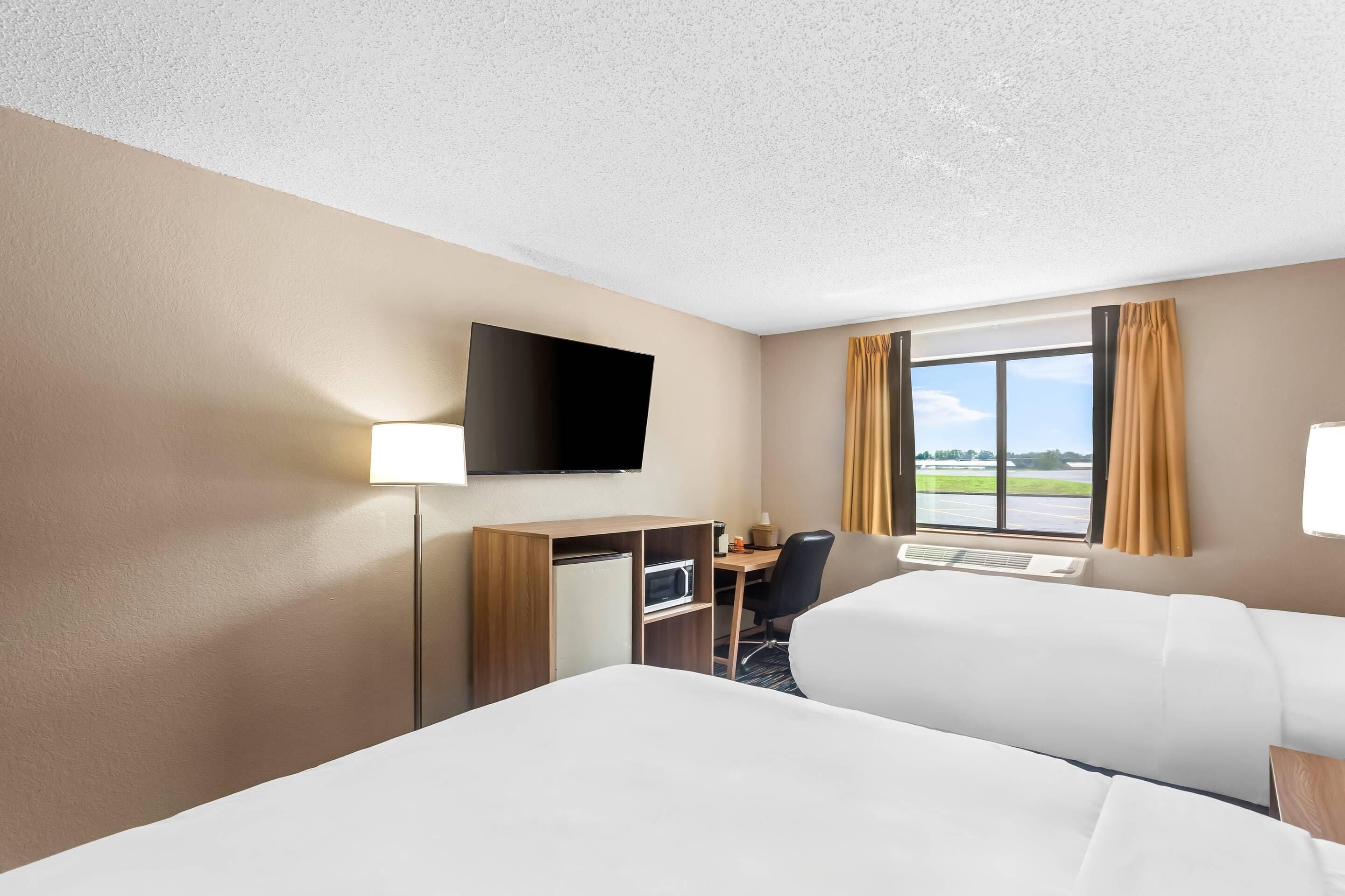 A hotel room with two beds, a desk with a chair, a flat-screen TV, and a window with curtains.