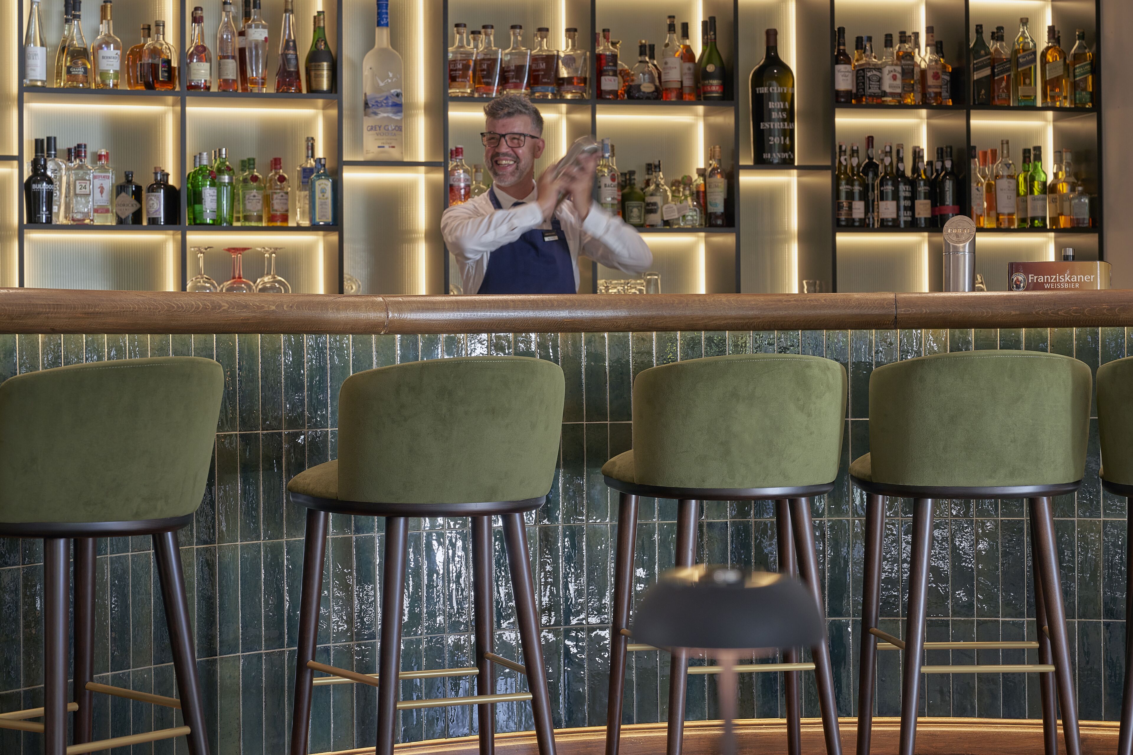 A bar area with a bartender, green bar stools, and shelves of liquor bottles.