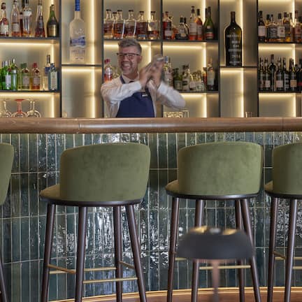 A bar area with a bartender, green bar stools, and shelves of liquor bottles.