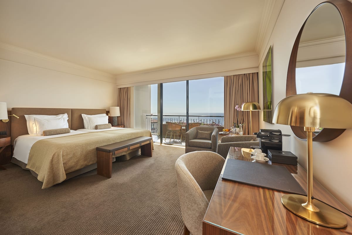 Habitación de hotel con cama, mesitas de noche, escritorio, silla, un espejo grande y vistas al mar a través de amplios ventanales.
