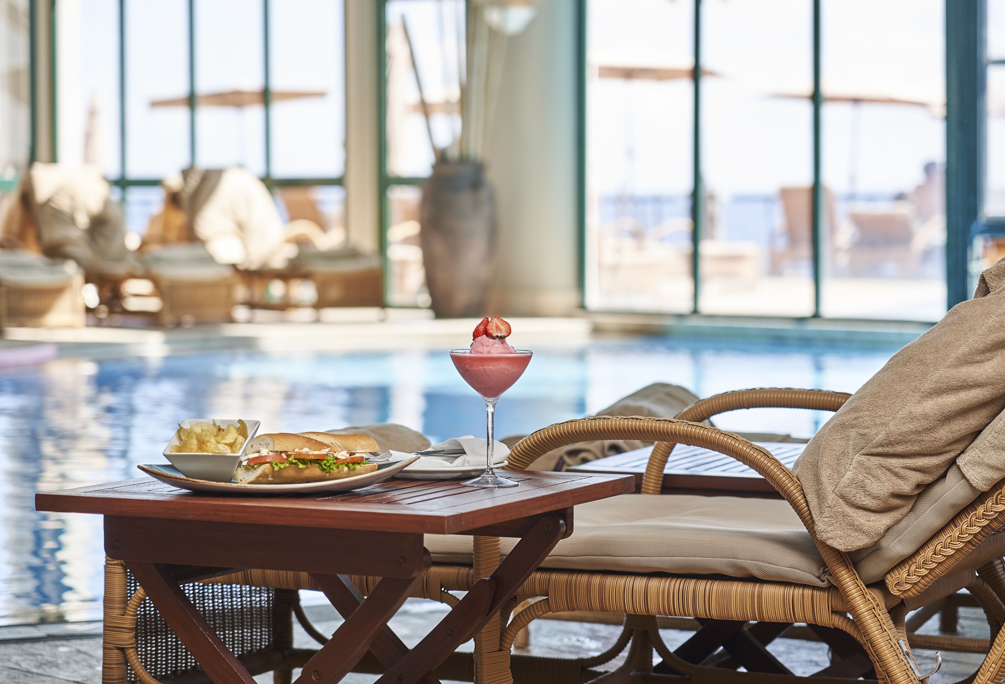 A dining table with a plate of food and a drink, a wicker chair, and a view of a pool area.