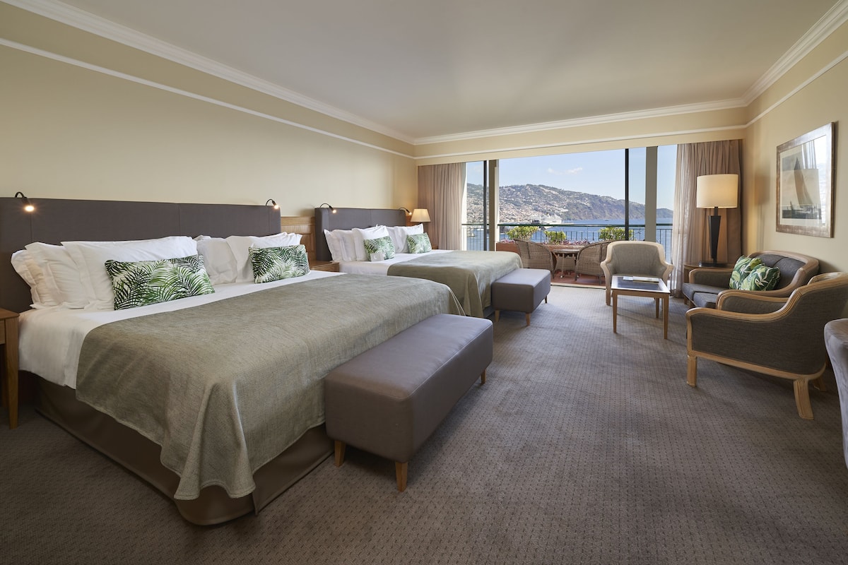Habitación de hotel con dos camas, zona de estar con mesa y sillas, y balcón con vista a las montañas.