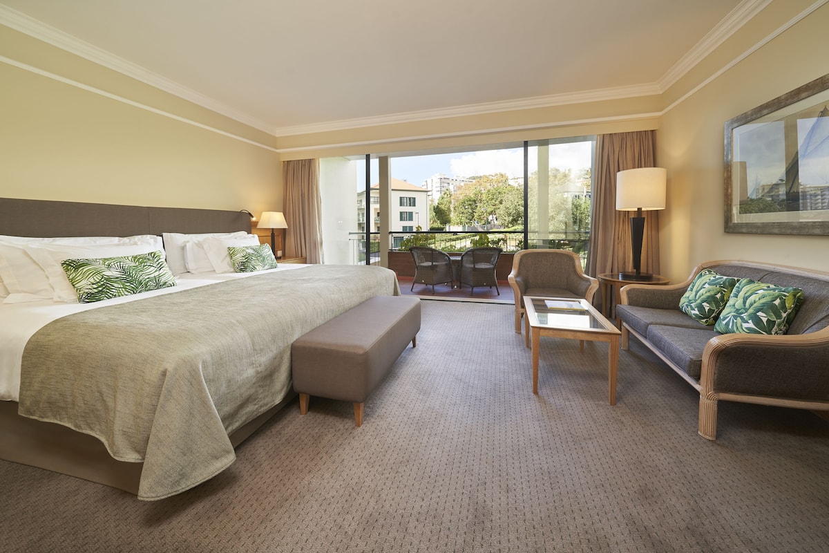 Habitación de hotel con una cama grande, un sofá, una mesita y una silla. Tiene un balcón con vistas a edificios y árboles.
