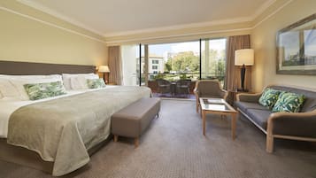 A hotel room with a large bed, a sofa, a small table, and a chair. There is a balcony with a view of buildings and trees.