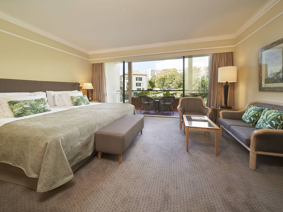 A hotel room with a large bed, a sofa, a small table, and a chair. There is a balcony with a view of buildings and trees.