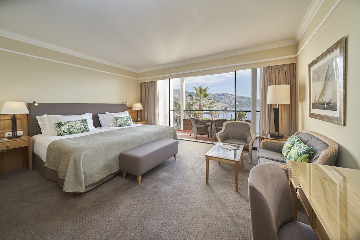 Habitación de hotel con una cama grande, un sofá, una mesita y un balcón con vistas al mar.