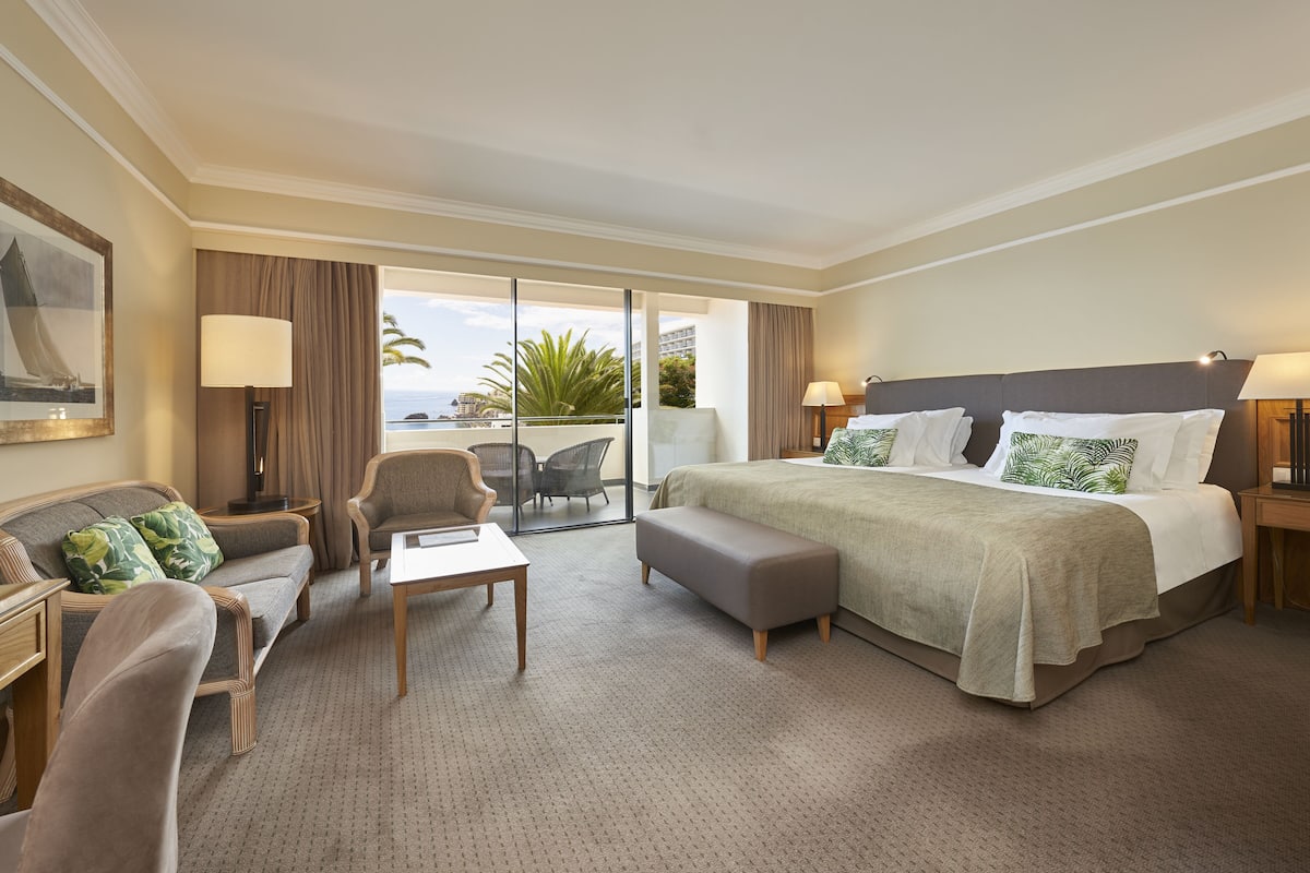 Habitación de hotel con cama, sofá, una mesita y una silla. Tiene un balcón con vistas al océano y palmeras.