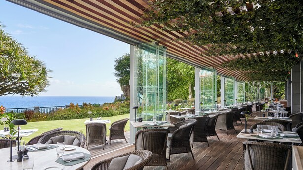 An outdoor dining area with wicker furniture, large glass windows, and a view of the ocean. - The Cliff Bay, by PortoBay (Funchal)