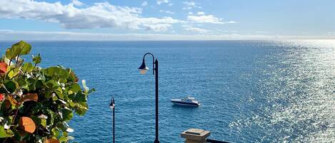 Una escalera conduce a un balcón con vista a un mar azul y despejado, donde se distingue un barco a lo lejos.