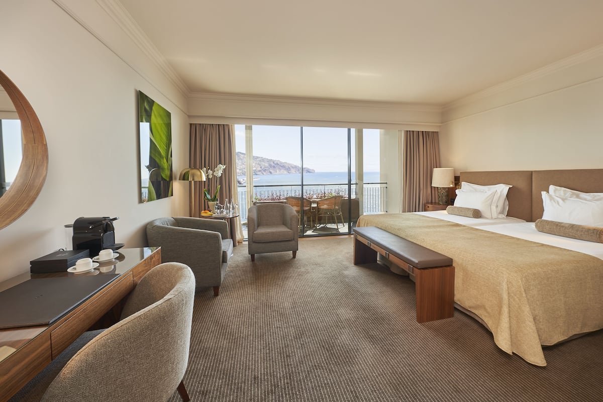Habitación de hotel con una cama grande, un escritorio con una computadora portátil, una silla, una mesita con una lámpara y vistas al mar.
