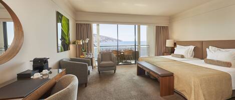 A hotel room with a large bed, a desk with a laptop, a chair, a small table with a lamp, and a view of the sea.