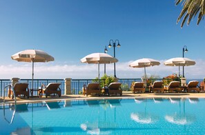 A poolside area with lounge chairs and umbrellas, a clear sky, and a view of the ocean.