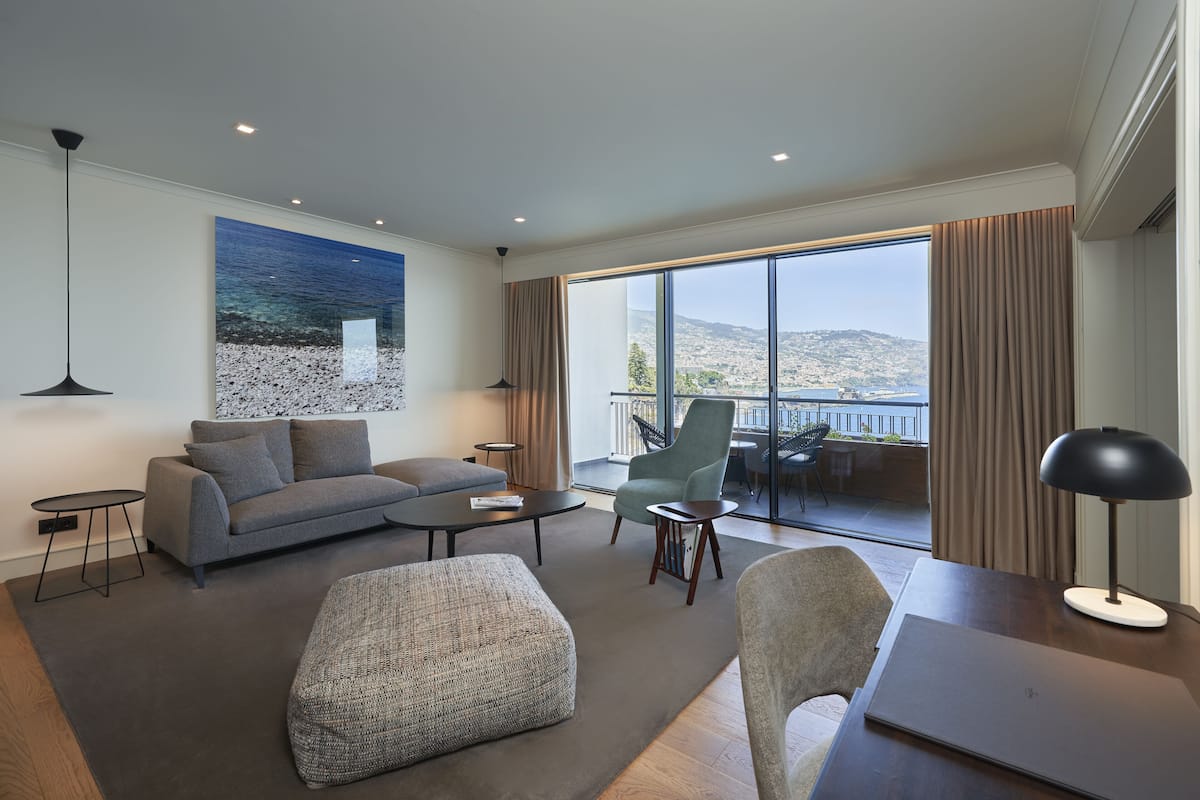 Una sala de estar moderna con un sofá, un reposapiés, una mesa de centro y un sillón. Un gran ventanal ofrece vistas a la ciudad y colinas.