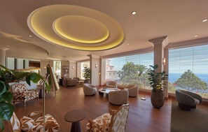 A spacious lobby with a modern design, featuring a central seating area with sofas and armchairs, large windows offering a view of the outdoors, and a ceiling with circular light fixtures.