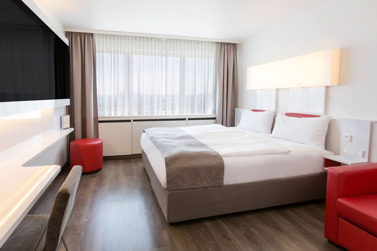 A modern hotel room with a large bed, a flat-screen TV, a red chair, and a window with sheer curtains.