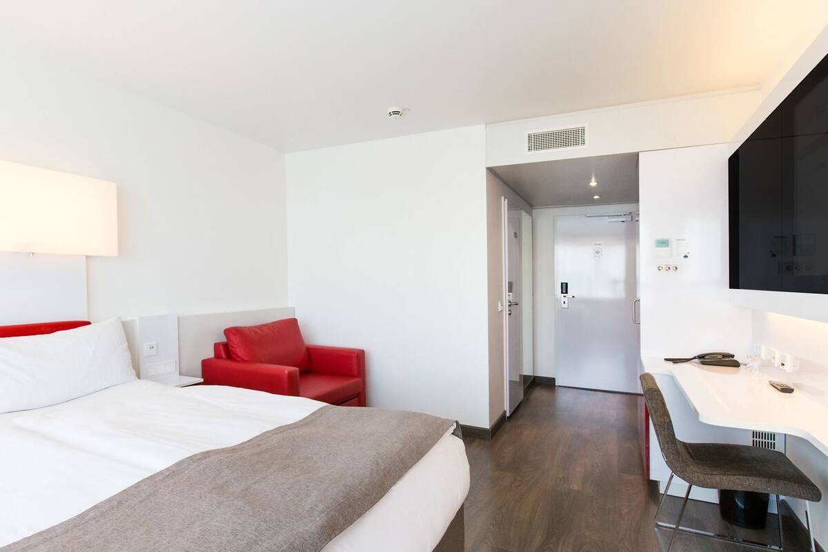 A modern hotel room with a bed, a red armchair, a desk, and a television.