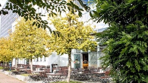 An outdoor seating area with tables and chairs, surrounded by trees and a building with large windows. - DORMERO Hotel Stuttgart (Stuttgart)