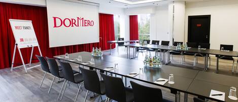 Un salón de conferencias con una larga mesa, sillas negras, una pizarra y una cortina roja.