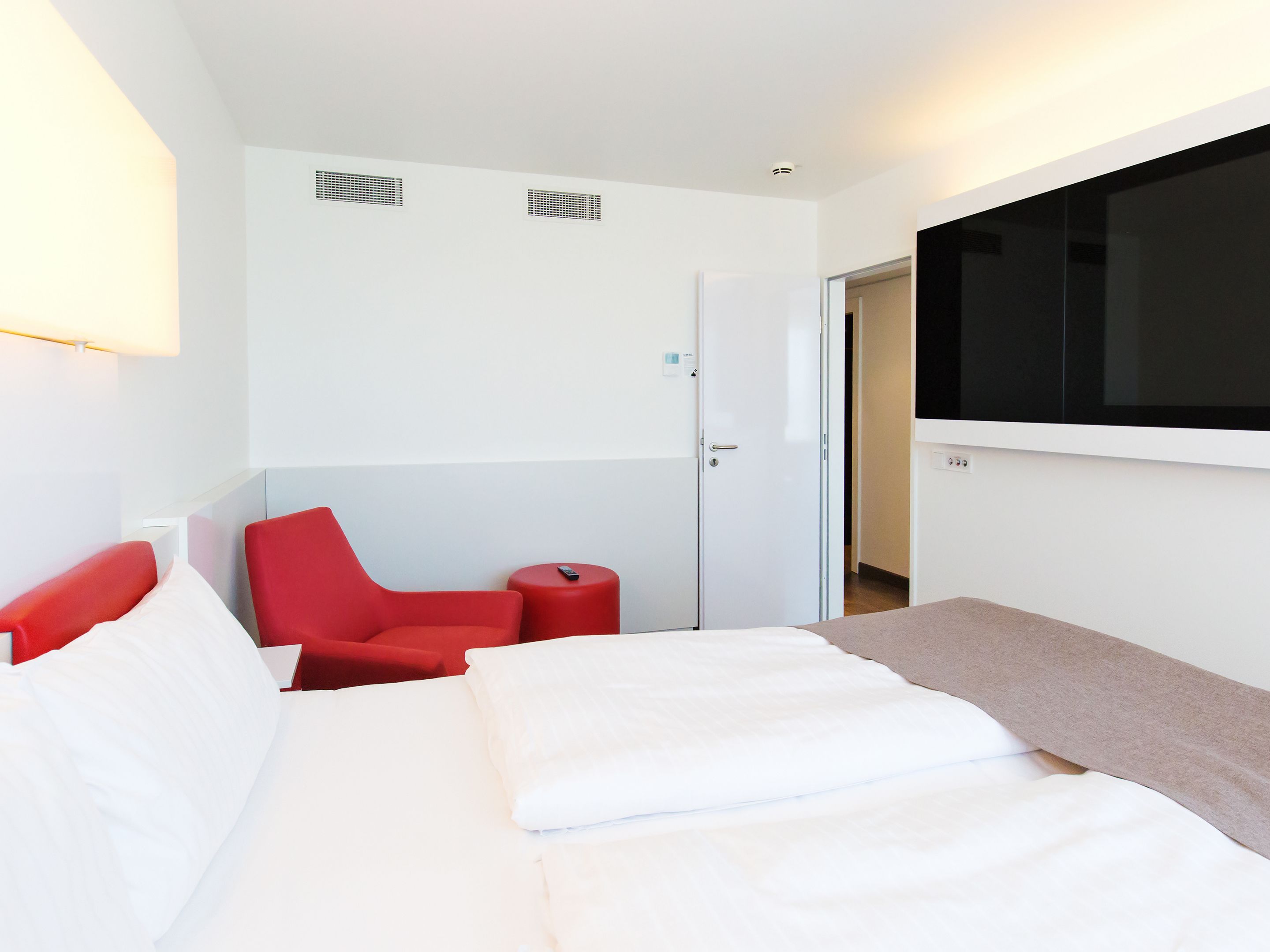Ein Hotelzimmer mit einem Bett, einem roten Sessel, einem kleinen Tisch und einem an der Wand montierten Fernseher.
