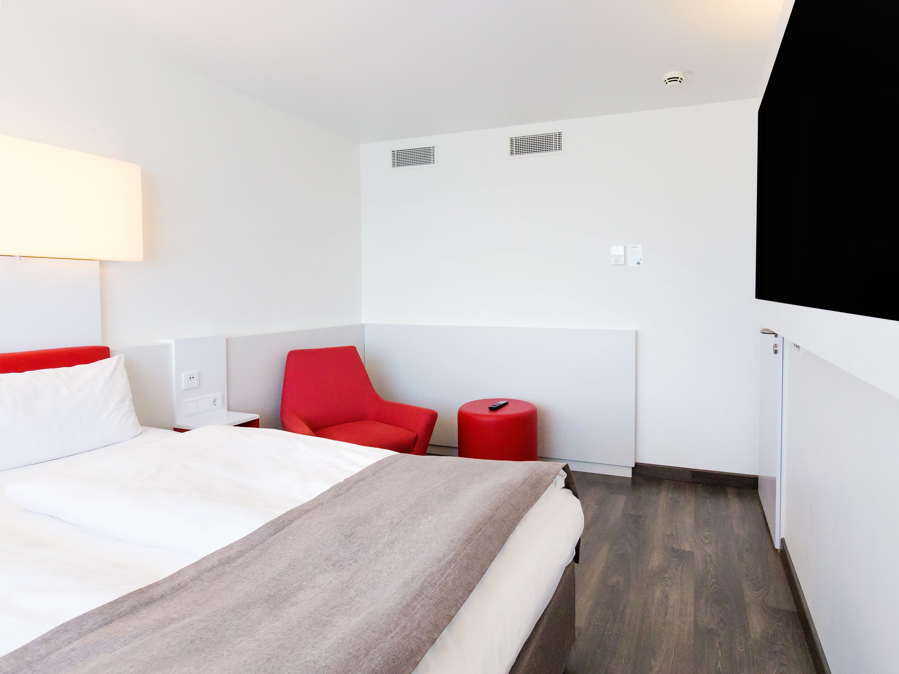 A hotel room with a bed, a red chair, a small table, and a red stool.
