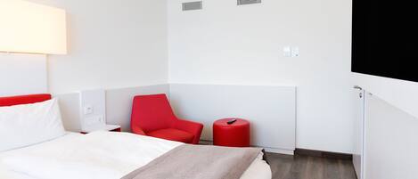 A hotel room with a bed, a red chair, a small table, and a red stool.