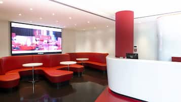 A modern reception area with red seating and white tables, a large screen displaying a room layout, and a red pillar.