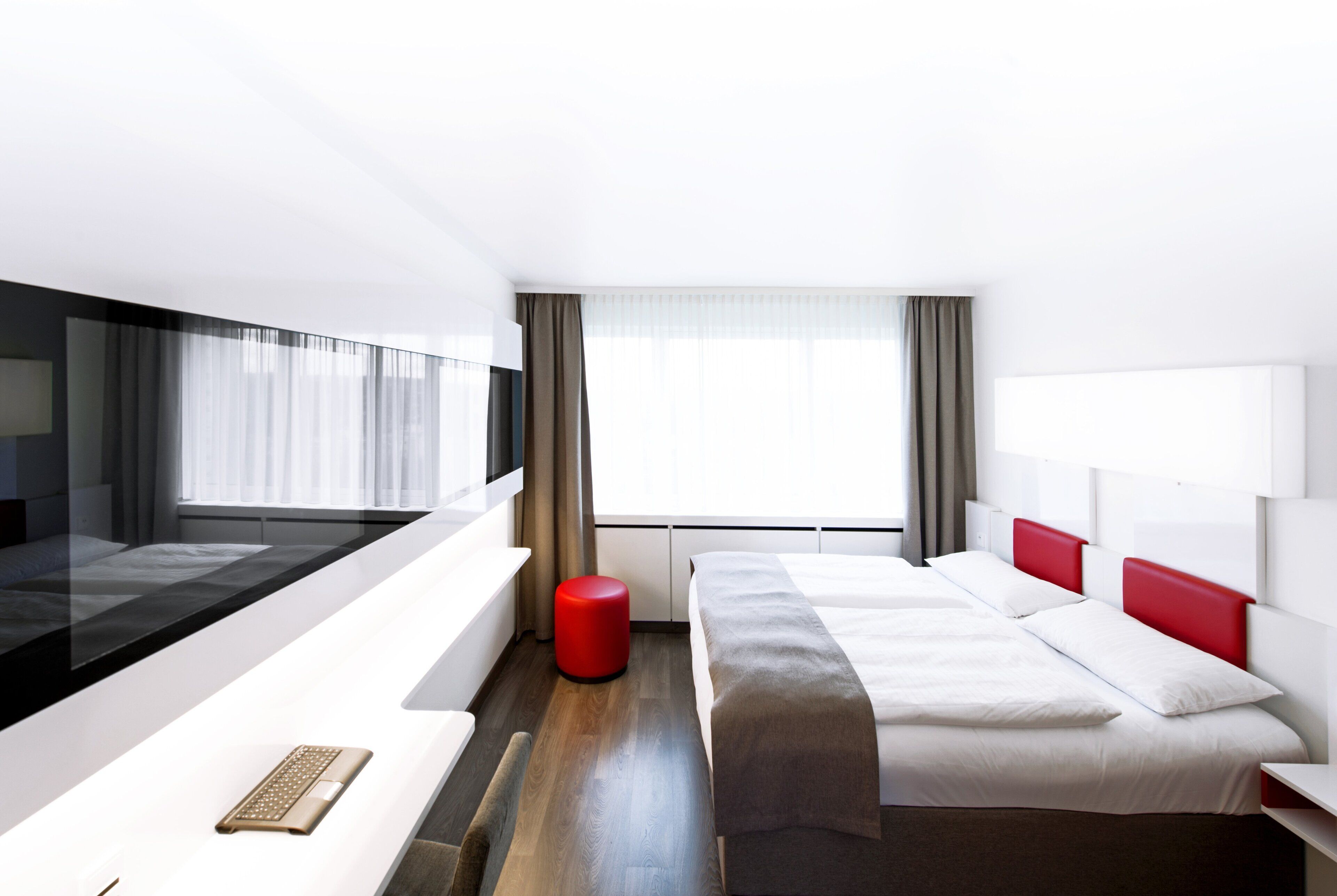 A modern hotel room with a large bed, a desk with a laptop, and a red ottoman.