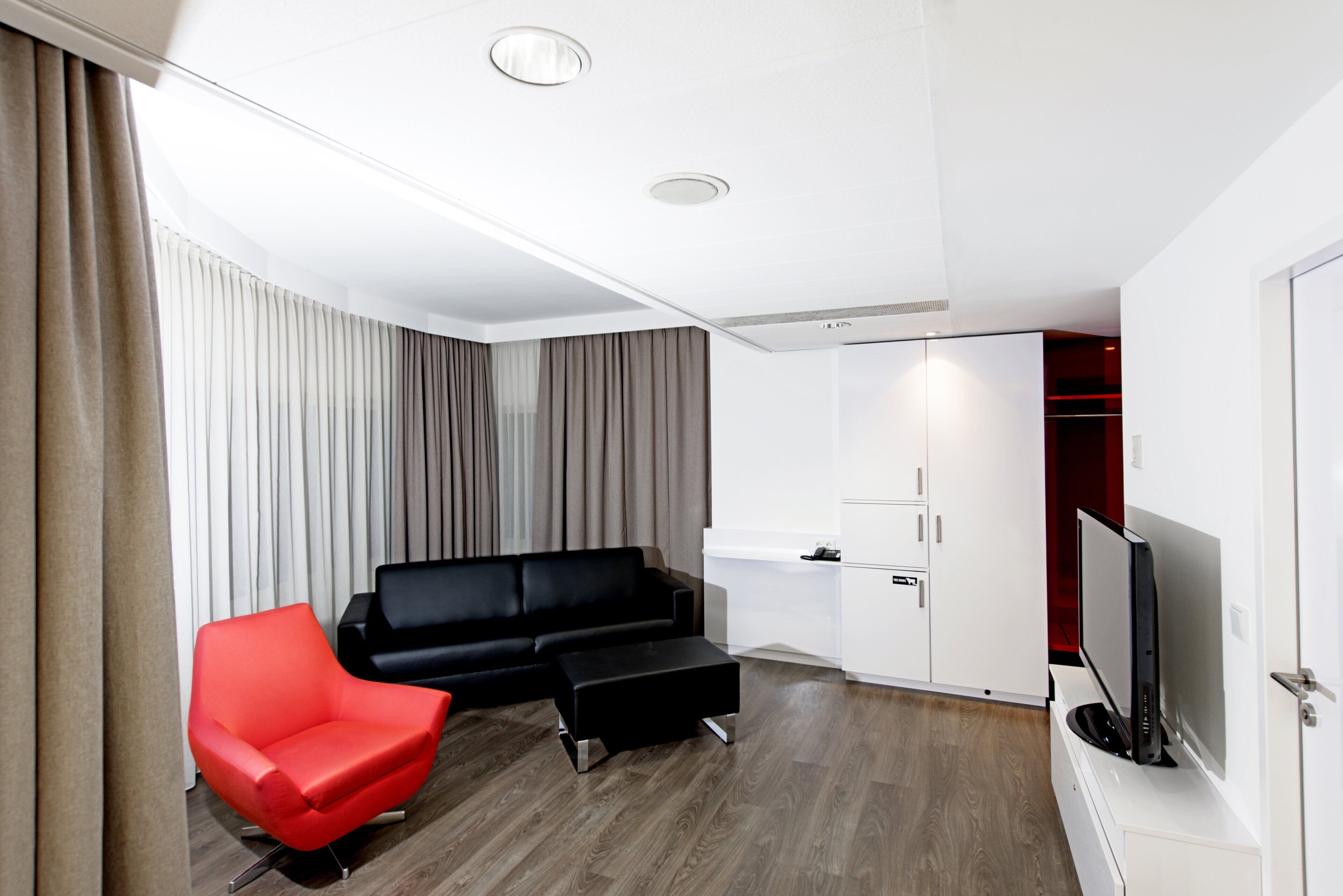 A modern living room with a black sofa, a red armchair, a black ottoman, a flat-screen TV, and a white cabinet.