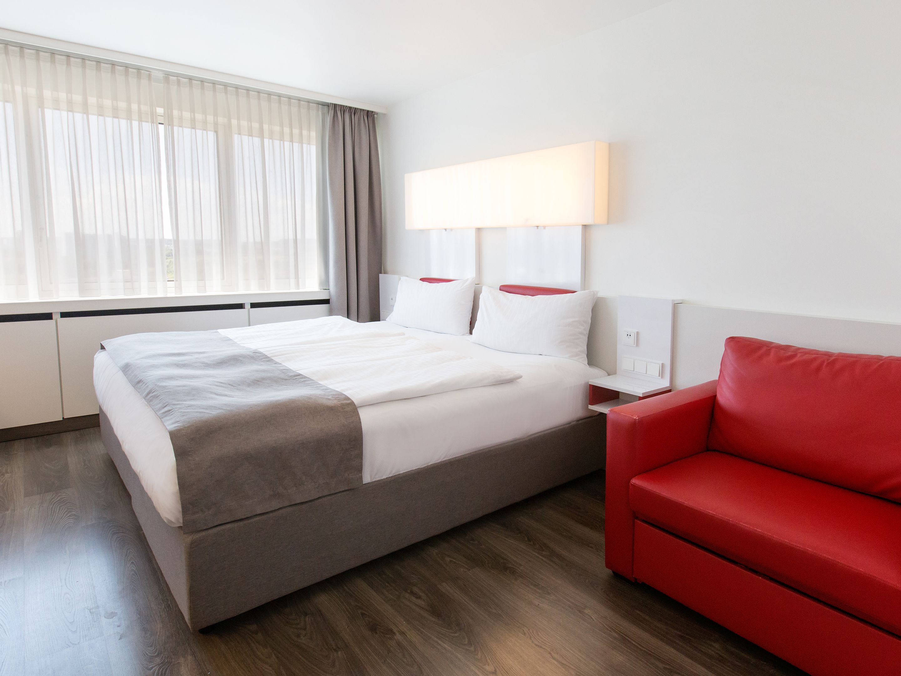 Ein Hotelzimmer mit einem Bett, einem roten Sofa, einem großen Fenster mit durchsichtigen Vorhängen und einer Wandlampe.