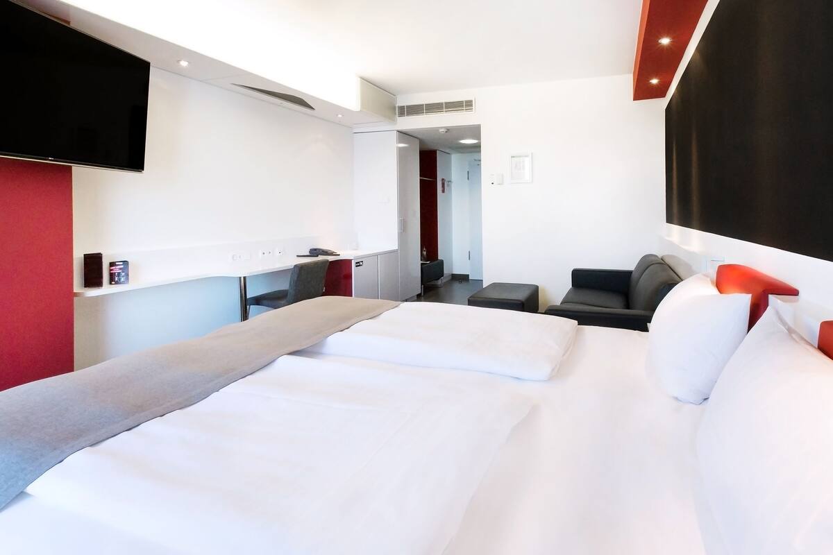 A modern hotel room with a large bed, a desk with a chair, a TV mounted on the wall, and a red accent wall.